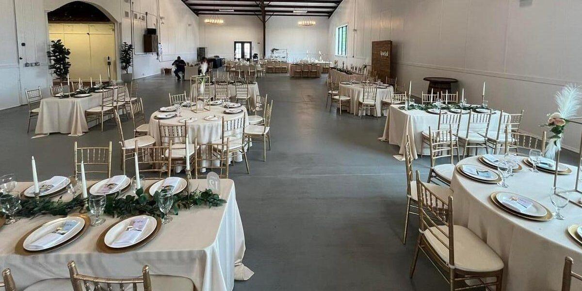 Round dinner tables set with white linens and gold chargers inside The Elegance indoor hall at The Dream Ranch, an event venue near Dallas