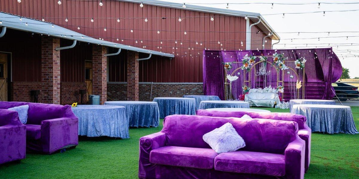 Purple velvet lounge sofas and blue-linen tables on the green turf cocktail area beside The Elegance barn at The Dream Ranch, a party venue near Dallas