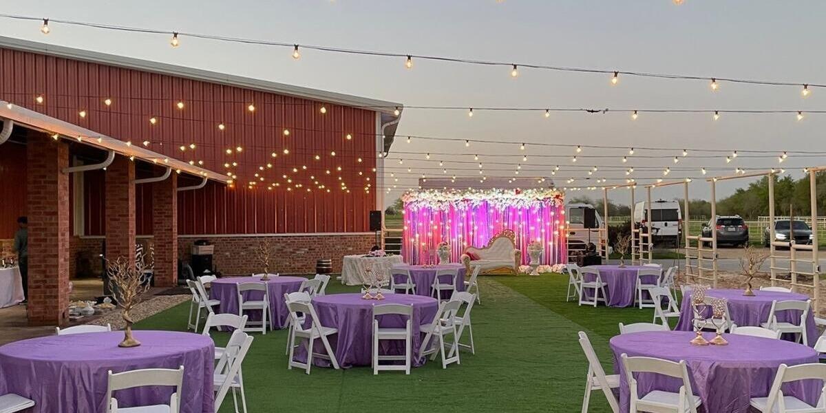 Round tables with purple linens and white chairs on a green lawn under string lights beside the red barn at The Dream Ranch near Dallas